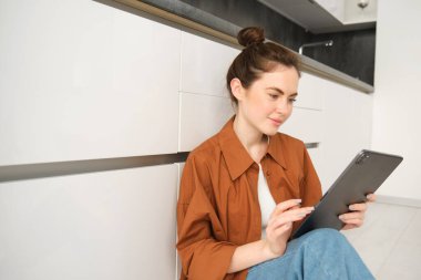 Evdeki güzel genç kadının portresi, mutfak zemininde oturuyor, dijital tablete bakıyor, cihaz kullanıyor, cihazda okuyordu..