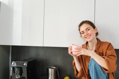 Güzel genç bir kadının portresi sabah kahvesinin tadını çıkarıyor, mutfakta oturup güneşli bir gün düşünürken çay içiyor..