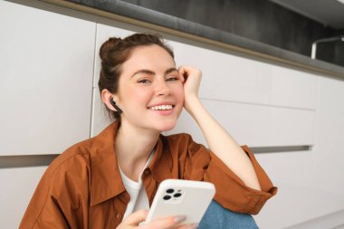 Akıllı telefonlu, mesajlaşan, cep telefonu kullanan, mutfak zemininde oturan, kablosuz kulaklıkla müzik dinleyen tasasız, güler yüzlü bir kadın..