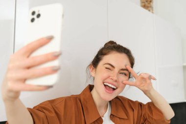 Pozitif, mutlu, cep telefonuyla selfie çeken, akıllı telefondaki fotoğraf için poz veren, barış işareti gösteren ve telefon kamerasına gülümseyen genç bir kadının yakın portresi..