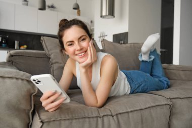 Oturma odasında kanepede dinlenen, elinde akıllı bir telefonla gülümseyen ve kameraya mutlu bakan bir kadının portresi. Yaşam tarzı kavramı