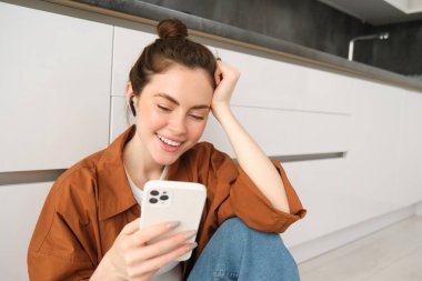 Kablosuz kulaklıklı genç bir kadının portresi, mutfak zemininde oturuyor, akıllı telefon kullanıyor, favori şarkılarla çalma listesi yapıyor, müzik dinliyor..