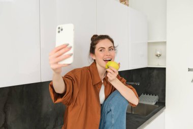 Mutlu kadın selfie çekmek için akıllı telefon kullanıyor, kamerada elma yiyor sosyal medya uygulamaları için video kaydediyor, mutfak tezgahında oturuyor..