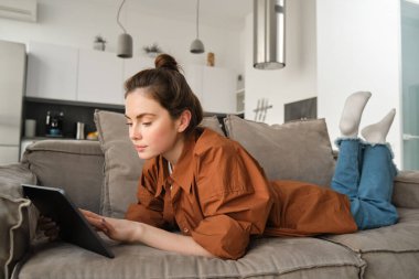 Hafta sonu ve yaşam tarzı konsepti. Genç bir kadın dijital tabletle kanepede uzanıyor, sosyal medyayı karıştırıyor, e-kitap okuyor ya da uygulamadan televizyon seyrediyor..