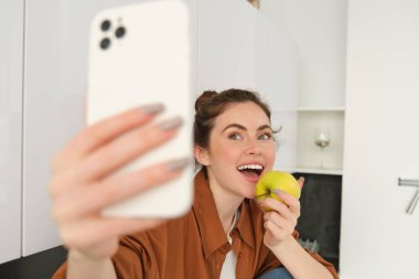 Güzel bir genç kadın, yaşam tarzı blogcusu kendini videoya kaydediyor, elma yerken selfie çekiyor, mutfak tezgahında oturuyor, akıllı telefon kullanıyor..