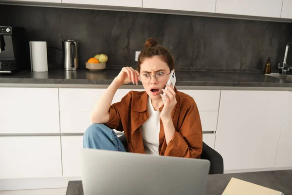 Portrait of woman with shocked face, receives concerning bad news over the phone conversation, sitting at home with laptop, working, talking to someone on telephone.