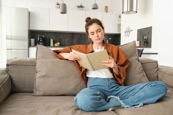 Image of woman relaxing on sofa, sitting and reading notebook, going through her notes in notepad.