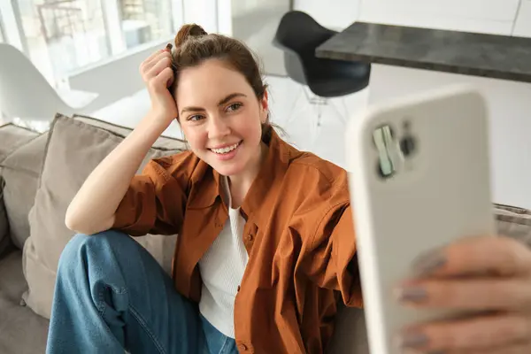 Tasasız, modern bir kadın, akıllı telefon uygulamasında gülümseyip selfie çekiyor, kanepesinde fotoğraf için poz veriyor, cep telefonuyla koltukta oturuyor..