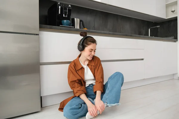 Cute modern girl on kitchen floor at home, listening music in wireless headphones, having fun.