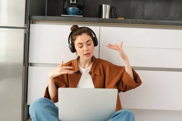 Portrait of woman listens to music in headphones, while working on laptop, sits at home on kitchen floor, making dance movements.