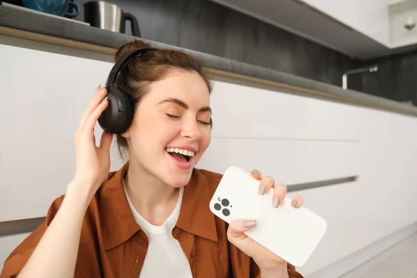 Happy woman listens to music, sings karaoke on smartphone app, sits on floor at home.