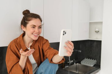 Telefon görüşmesi sırasında gülen ve gülümseyen genç bir kadının portresi, arkadaşlarıyla görüntülü sohbetler, mutfak tezgahında oturuyor ve akıllı telefon uygulaması kullanan biriyle konuşuyor..