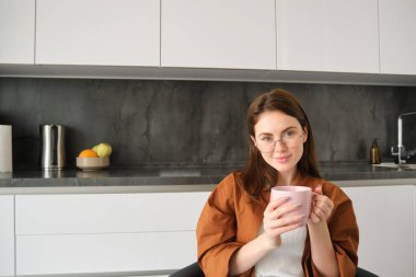 Huzur içinde kahve içen genç bir kadının portresi, evinde oturmuş, elinde bitki çayıyla fincanı tutuyor, gözlük takıyor, mutfakta dinleniyor..