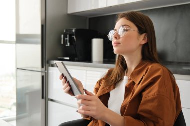 Genç bir kadının portresi mutfakta, elinde dijital tablet, gözlük takıyor, düşünceli görünüyor, proje üzerinde çalışıyor, ders çalışıyor..
