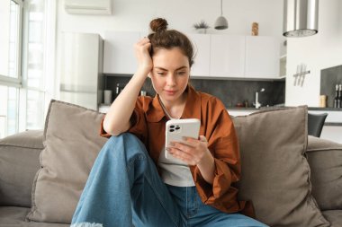 Cep telefonuyla kitap okuyan genç bir kadının portresi, akıllı telefondaki sosyal medya uygulaması, oturma odasındaki kanepede günlük kıyafetlerle oturması..