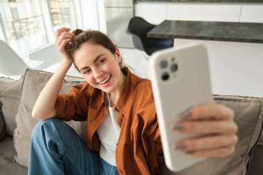 Güzel, mutlu genç kadın modelin portresi, akıllı telefonlu koltukta oturuyor, cep telefonuyla selfie çekiyor, gülen surat yapıyor..