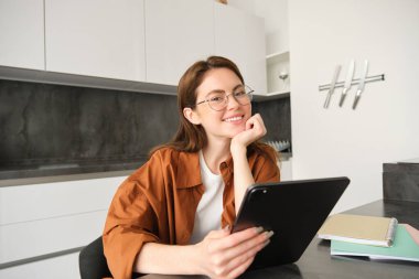 E-öğrenme ve uzak iş yeri konsepti. Gözlüklü genç bir kadın, mutfakta okuyan bir öğrenci, dijital tablet okuyor, projesine bakıyor, online toplantıya bağlanıyor..