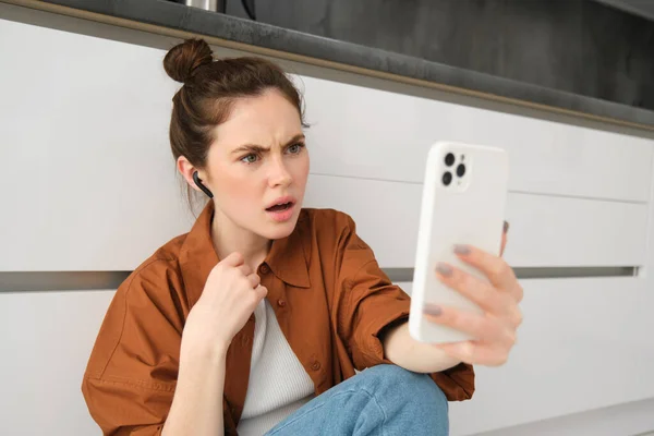 Portrait of woman with shocked, frustrated face, looking at smartphone screen, hear bad news over the phone, talking with wireless headphones.