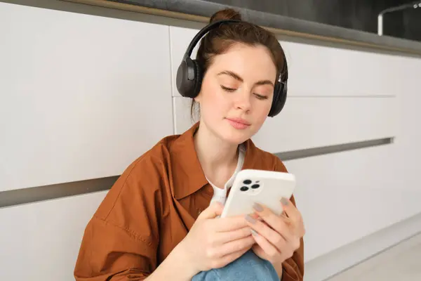 Young modern woman sending a message on smartphone, sits at home in wireless headphones, listens music, watches video on mobile app.