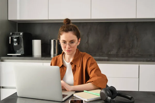 Work and life balance concept. Young woman in glasses, working from home, typing on laptop, student doing homework on computer, sitting in kitchen, freelancing.