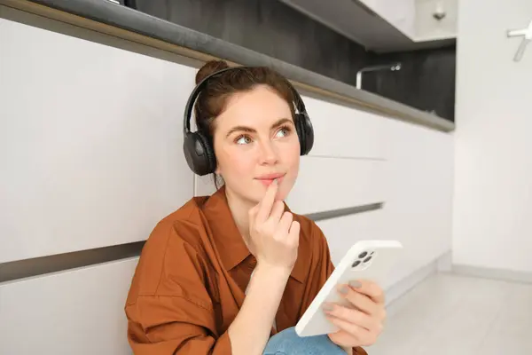 Close up of young woman sits on floor at home, listens music in headphones, holds smartphone, looks thoughtful, orders online on mobile phone app.