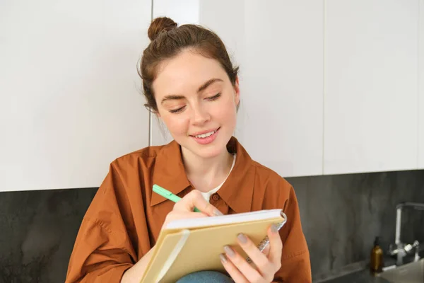 Portrait of cute young woman with notebook, sitting in the kitchen and writing her to do list, making tasks for week, puts notes in schedule or planner.