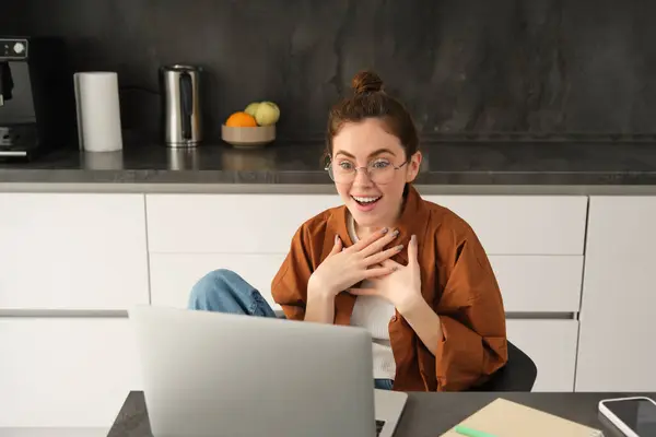 Portrait of beautiful woman in glasses, sits at home in kitchen, looks at computer screen with amazed, surprised face, reacts to big great news seeing it on laptop.