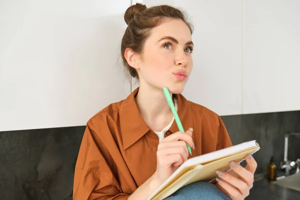 Close up of thoughtful young woman with notebook, looking up with thinking face, writing down her ideas or thoughts in planner, makes list of groceries.