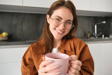 Gözlüklü güzel bir kızın portresi, elinde sıcak bir bardak içki, evde çay ya da kahve içip kameraya gülümsüyor..