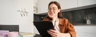 E-öğrenme ve uzak iş yeri konsepti. Gözlüklü genç bir kadın, mutfakta okuyan bir öğrenci, dijital tablet okuyor, projesine bakıyor, online toplantıya bağlanıyor..