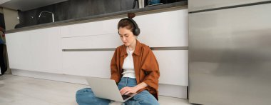 Serbest çalışan genç bir kadın, mutfakta dizüstü bilgisayarla oturan, kulaklık takan, bilgisayar başında yazan bir öğrenci..