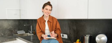 Şık bir genç kadının portresi, mutfakta oturur ve notlar alır, yemek tarifini yazar, ev işleriyle listesini kontrol eder, defterine karalama veya çizim yapar..