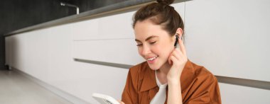 Akıllı telefondan kulaklıkla video izlemekten hoşlanan bir kadının portresi, müzik dinleyen ve cep telefonu kullanan bir kız, mutfak zemininde oturuyor..