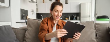 Dijital tablette video oyunu oynayan genç bir kadının portresi, heyecanlı görünüyor, aleti eğiyor ve ekrana bakıyor, oturma odasında kanepede oturuyor..