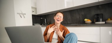 Evden çalışmak, uzaktan iş kavramı. Genç bir kadın mutfakta oturuyor, dizüstü bilgisayar kullanıyor, görüntülü sohbete katılıyor, çevrimiçi dersler veriyor, bilgisayarda müşteriyle konuşuyor..