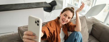 Güzel mutlu kızın portresi kanepede selfie çekiyor. Kanepesinde poz veren, sosyal medya için fotoğraf çeken bir kadın akıllı telefonuyla elini uzatıyor..