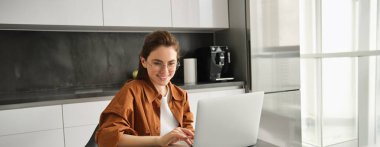 Serbest çalışan genç kadın portresi, dijital göçebe evde çalışıyor, dizüstü bilgisayar kullanıyor ve gözlük takıyor, çalışma alanını mutfakta kuruyor..