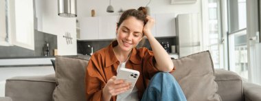 Kanepede oturan mutlu esmer kadının portresi akıllı telefonlu, bildirim okuyan, cep telefonuyla mesajlaşan, evdeki koltukta dinlenen, cihaz ekranına bakarken gülümseyen.