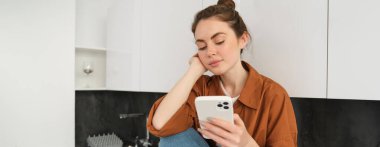 Modern stil kızı görüntüsü mutfak tezgahında telefonuyla oturuyor, cep telefonuna bakıyor, mesajları kontrol ediyor, akıllı telefondaki sosyal medya uygulamasını kaydırıyor..