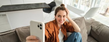 Şık, modern genç bir kadının portresi akıllı telefonuyla selfie çekiyor, koltukta cep telefonuyla oturuyor, yaşam tarzı blogu için fotoğraf çekiyor..