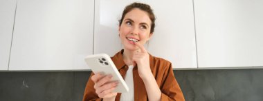 Heyecanlı genç bir kadının portresi, gülümseyen ve düşünen bir yüz, alışveriş uygulaması kullanırken, akıllı telefon tutarken.