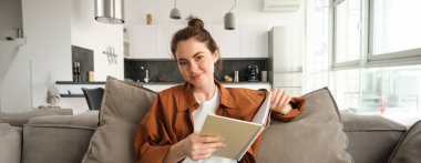 Güzel genç bir kız öğrenci, kanepede oturuyor, kanepede dinleniyor, defter okuyor ve gülümsüyor..