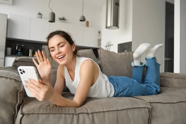Happy young woman video chatting with friends, using smartphone app, waving hand at smartphone screen and saying hello.