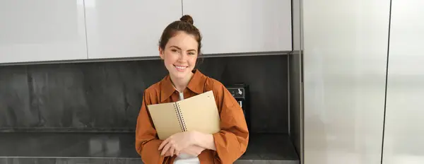 Stock image Portrait of beautiful modern woman, standing in the kitchen, flipping recipe book, holding notebook and smiling, reading notes.