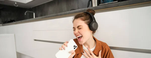 Happy woman listens to music, sings karaoke on smartphone app, sits on floor at home.