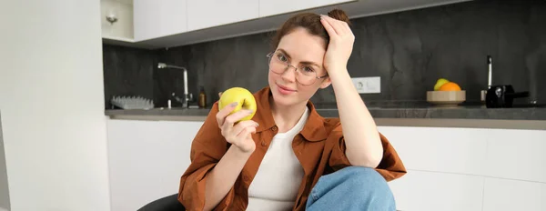 Portrait of smiling, modern woman in glasses, sitting at home and eating green apple, having a lunch break, biting fruit.