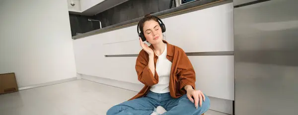 stock image Technology concept. Young smiling brunette woman in wireless headphones, listens to music, sits on kitchen floor.