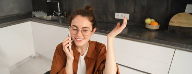 Mutfakta gözlükleriyle oturan, cep telefonuyla konuşurken el kol hareketi yapan, telefonlara cevap veren, akıllı telefon kullanan mutlu ve arkadaş canlısı genç bir kadının portresi..