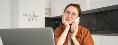 Evden çalışan güzel gülümseyen bir kadının portresi, cep telefonuyla konuşan, müşteriyi arayan, serbest çalışan bir iş kadını, dizüstü bilgisayar ve akıllı telefon kullanarak mutfakta iş yeri kuruyor..