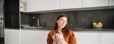 Huzur içinde kahve içen genç bir kadının portresi, evinde oturmuş, elinde bitki çayıyla fincanı tutuyor, gözlük takıyor, mutfakta dinleniyor..